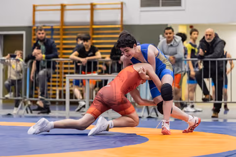 Two young wrestlers battle on the mat; blue singlet athlete drives forward aggressively while red singlet opponent braces low.