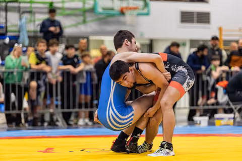 Young wrestler in blue singlet drives forward executing a double-leg takedown against black singlet opponent on yellow mat