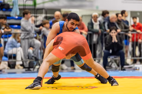 Young wrestler in blue singlet smiles while controlling opponent in red during match on yellow mat, crowd watching.