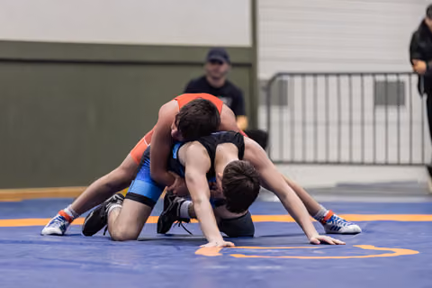 Two young wrestlers in red and blue singlets battle on the mat, one gaining control from behind during intense match.