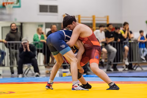 Two young wrestlers locked in a grapple on yellow mats, blue vs red singlet, crowd watching intently behind barriers.