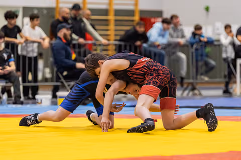 Two young wrestlers battle for control on the mat, one in blue pinned low by opponent in black and red singlet.