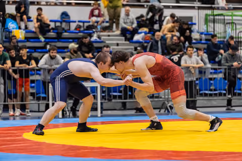 Two wrestlers in navy and red singlets lock hands in an intense face-off on a colorful mat before a packed crowd.