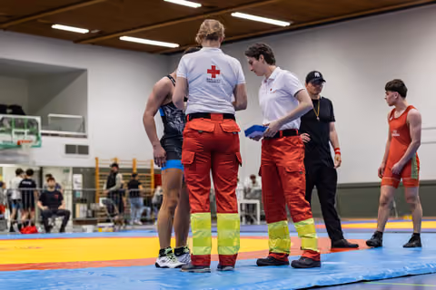 Two Red Cross paramedics consult with a wrestler on the mat during a break, while a referee and athlete look on.
