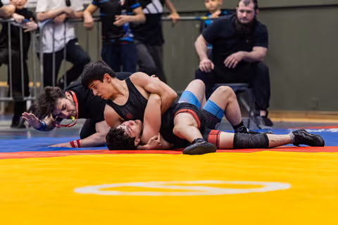 Young wrestler pins opponent on mat, referee watches closely as crowd looks on in tense indoor bout.