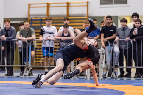 Young wrestler executes a dramatic throw, lifting opponent overhead on a blue mat as spectators watch intently.