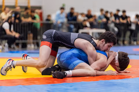Wrestler in dark singlet pins opponent in blue to the red mat, crowd watching in background.
