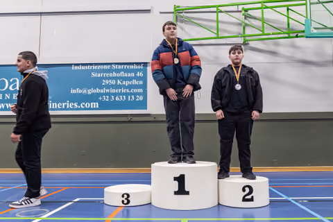 Two boys stand on podium positions 1 and 2 wearing medals, while a third walks past position 3 in an indoor sports hall.