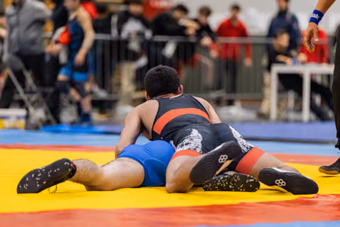 Wrestler in black singlet pins opponent in blue to the yellow mat during an indoor tournament match.