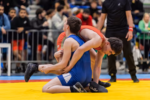 Wrestler in red lifts opponent in blue from behind on yellow mat, crowd watching in background.