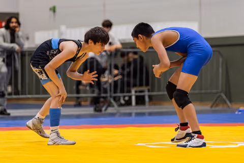Two young wrestlers crouch in a tense standoff on a yellow mat, focused and ready to engage in a gym.