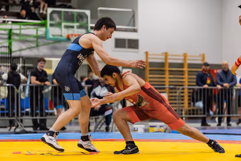 Two young wrestlers engage in a tense standoff on a yellow mat, one in navy blue, one in red, both crouching low.