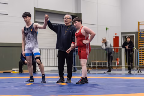 Referee raises the arm of a wrestler in blue singlet declaring victory, while opponent in red stands nearby.