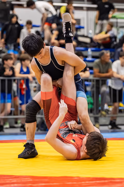 Young wrestler in navy singlet lifts and throws opponent in red onto the yellow mat during a tense bout.