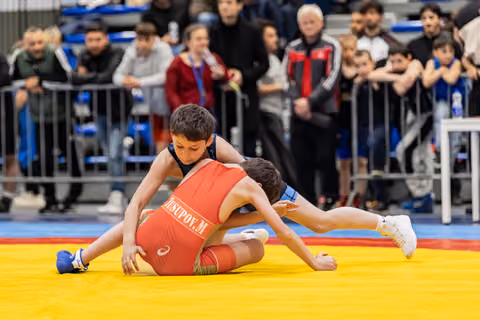 Boy in blue singlet controls opponent Yusupov M in red on yellow mat, crowd watching intently in background.