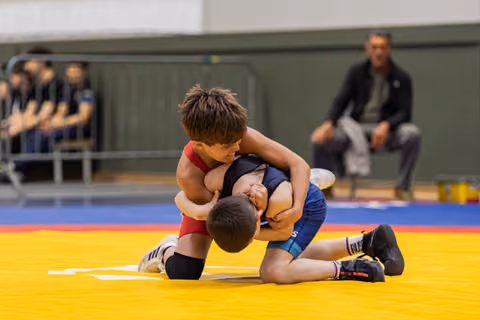 Two young boys wrestle on a yellow mat, one in red dominating with a headlock grip during an indoor tournament.
