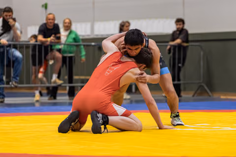 Two young wrestlers grapple intensely on a yellow mat, one in red kneeling as the other dominates from above.