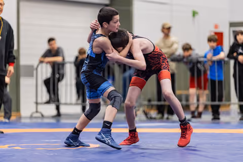 Two young wrestlers locked in a clinch on a blue mat, both focused and straining as they grapple for control.