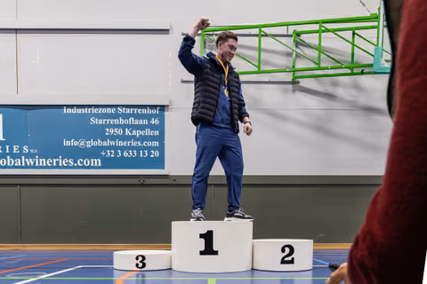 Young male athlete raises fist in triumph on 1st place podium, wearing a gold medal, smiling in an indoor sports hall.