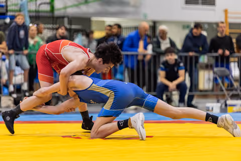 Wrestler in red singlet aggressively drives blue-clad opponent down onto the yellow mat during an intense match.