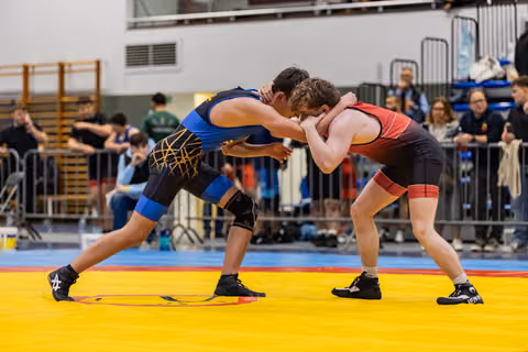 Two young male wrestlers grip each other in a standing clinch, both low and intense on a yellow competition mat.