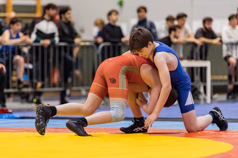 Two young wrestlers in red and blue singlets locked in a ground grapple, intense focus during an indoor tournament.