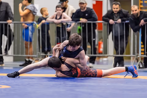 Two young wrestlers battle on the mat, one pinning the other down, with coaches and spectators watching intently behind barriers.
