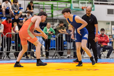 Two young wrestlers in orange and blue singlets crouch in a tense standoff on a yellow competition mat, referee watching closely.