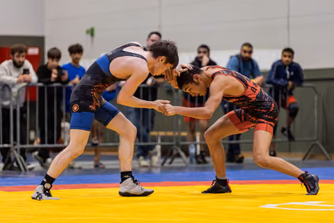 Two male wrestlers in intense grappling standoff on yellow mat, spectators watching behind barriers in indoor arena.