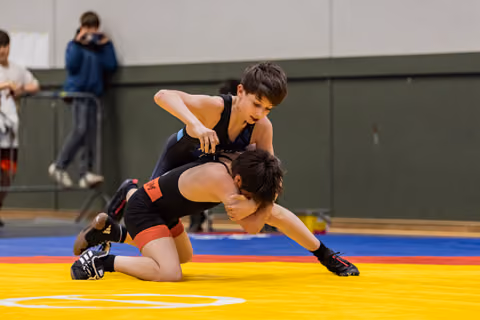 Young wrestler in navy singlet dominates opponent on colorful mat, intense focus during match.