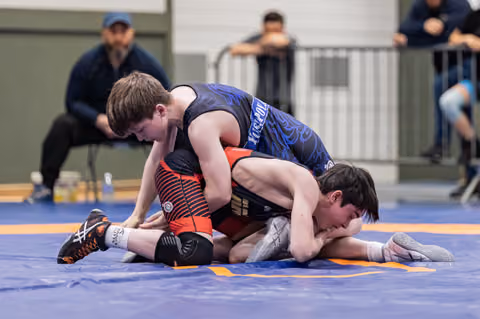 Young wrestler in blue singlet controls opponent on blue mat, spectators watching in background during indoor meet.