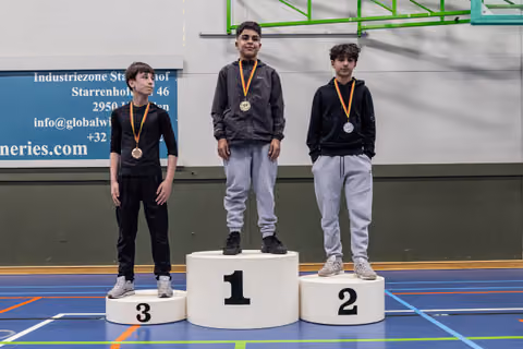 Three boys stand on a podium with medals; 1st place stands tallest, 3rd looks downcast, indoors.