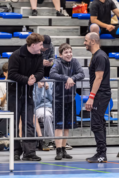 Bald coach and young teen share a laugh with an older youth beside a barrier at an indoor sports event.