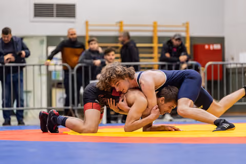 Curly-haired wrestler in black controls opponent in navy singlet on colorful competition mat, spectators watching