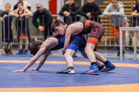 Teen wrestler in red-black singlet controls opponent on blue mat, spectators watching intently in background.
