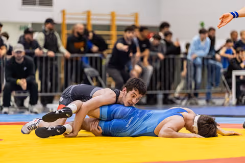 Wrestler in black singlet pins opponent in blue to the yellow mat, crowd watching intently in background.