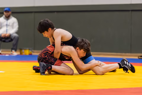 Two young wrestlers compete on a colorful mat, one in black pinning the other in red from above with focused intensity.