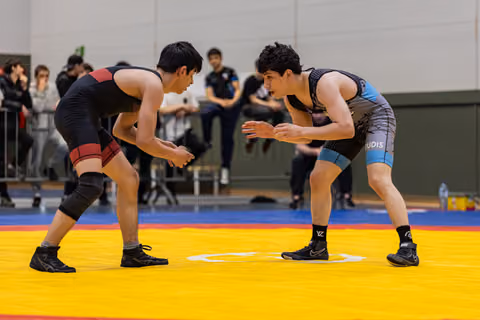 Two young male wrestlers in crouched stances face each other intently on a yellow competition mat, spectators watching behind.