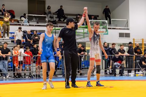 Referee raises the white-singlet wrestler's arm in victory while the blue-singlet opponent walks away deflated.
