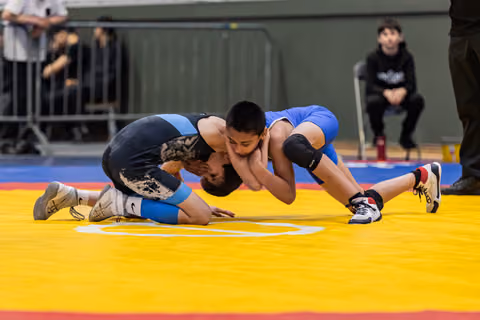Two young wrestlers in blue and black singlets locked in a ground clinch on a yellow competition mat, focused intensity.