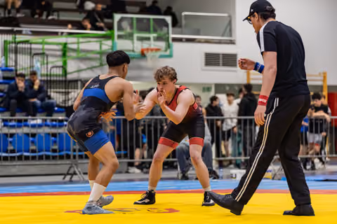Two teen wrestlers face off in tense stance on yellow mat, referee watching closely in indoor sports hall.