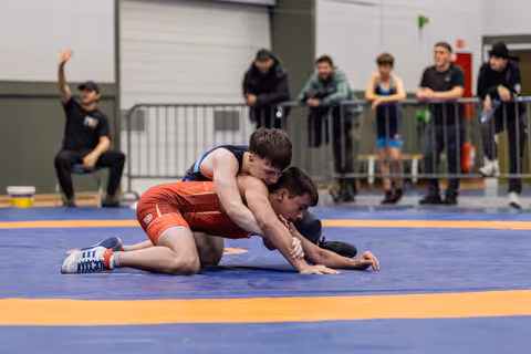 Young wrestler in blue singlet controls opponent in red on the mat, referee watches closely in indoor sports hall.