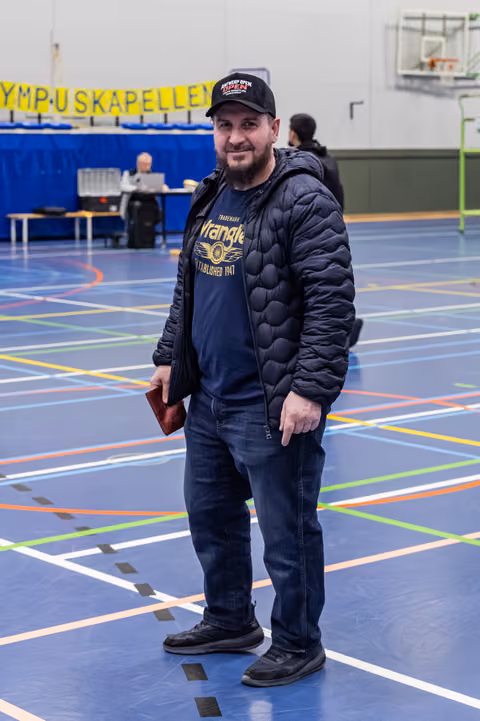 Bearded man in Wrangler tee and black jacket stands relaxed on a multi-sport gymnasium floor, smiling at camera.