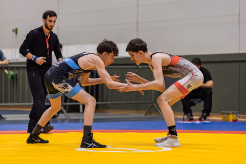 Two teen wrestlers crouch in tense standoff, hands reaching toward each other on a yellow mat, referee watching closely.