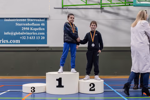 Two boys shake hands on a winner's podium, both smiling with medals around their necks in an indoor sports hall.