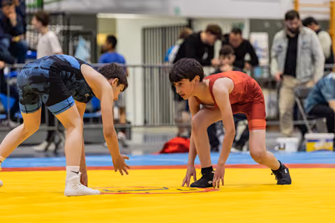 Two young wrestlers in blue and red singlets crouch in a tense face-off stance on a yellow competition mat.