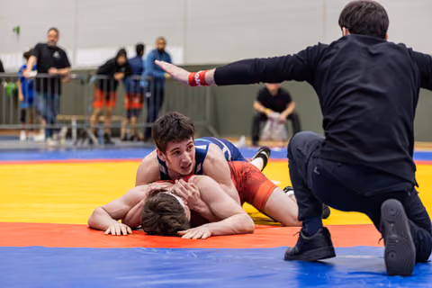 Young wrestler in blue pins opponent face-down on mat as referee kneels and raises hand to signal.