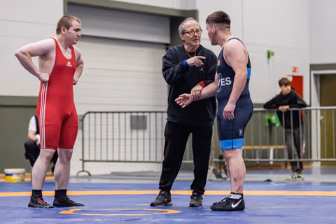 Coach gestures instructively between red and blue singlet wrestlers on competition mat, both listening attentively.
