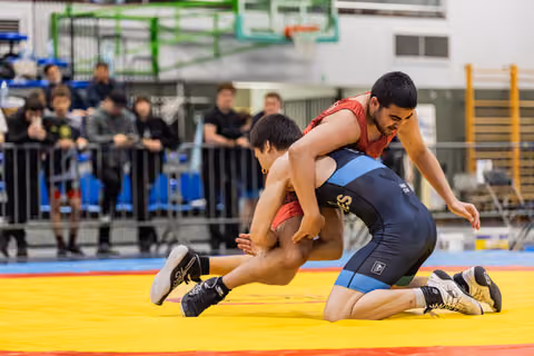 Wrestler in red singlet drives opponent down onto the yellow mat during an intense indoor match, crowd watching.