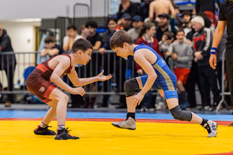 Two young wrestlers in red and blue singlets face off in crouched stances, eyes locked, crowd watching behind barriers.